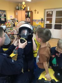 Policjant pomaga zakładać maskę przeciwgazową chłopcu ubranemu w mundur policyjny i kask.