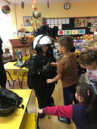 Chłopiec ubrany w kamizelkę kamienioodporną i kask policyjny. Jest w towarzystwie innych dzieci i policjanta.
